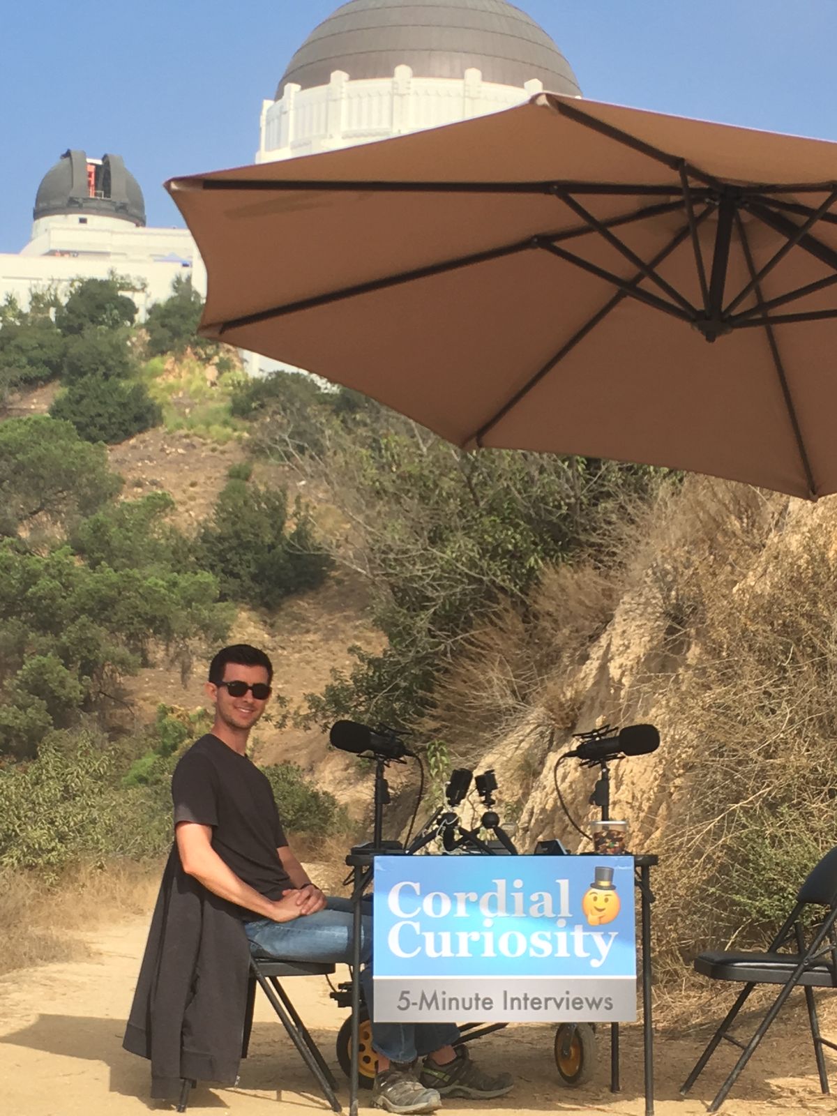 A man sitting at a Cordial Curiosity 5-Minute Interviews table on a hillside path with the Griffith Observatory behind him
