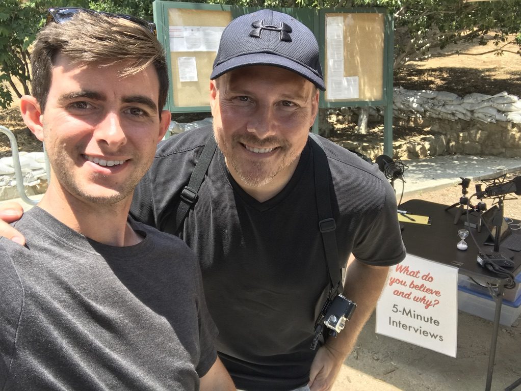 Two men smiling in a selfie in front of a table with a sign reading What do you believe and why? 5-Minute Interviews