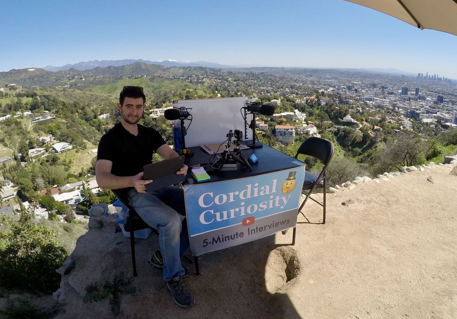 Reid sitting at a Cordial Curiosity 5-Minute Interviews table on a hilltop with the Hollywood Sign and Los Angeles behind him