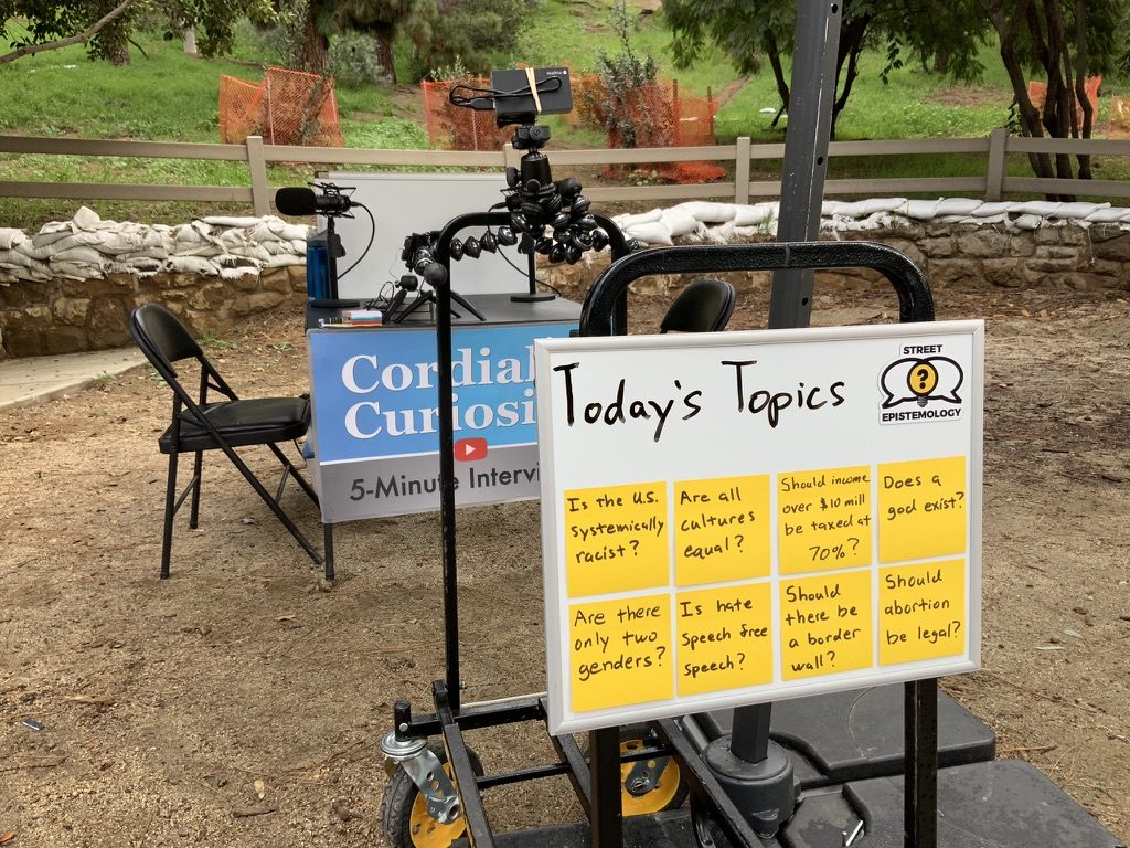 A Today's Topics whiteboard with sticky-note questions next to a Cordial Curiosity 5-Minute Interviews table in a park